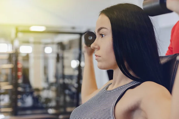 Una joven que usa ropa deportiva en un gimnasio realiza ejercicios de mancuerna, el entrenador la ayuda — Foto de Stock