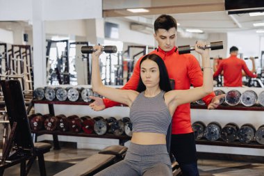 Bir genç kız bir spor salonunda spor giyim giyiyor halter alıştırmalar gerçekleştirir, Koç ona yardımcı olur