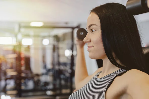 Una joven que usa ropa deportiva en un gimnasio realiza ejercicios de mancuerna, el entrenador la ayuda — Foto de Stock