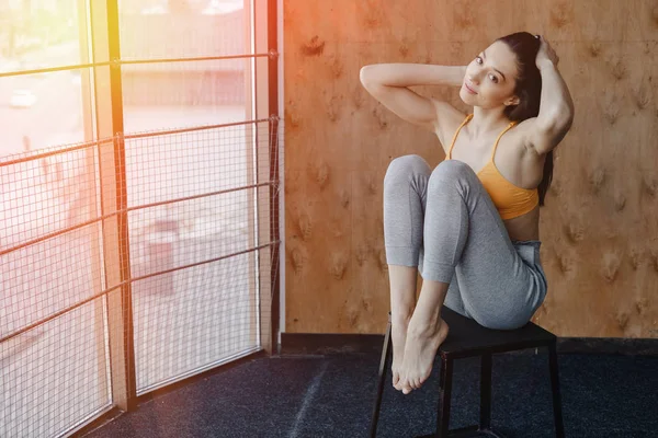 Joven chica atractiva fitness sentada en la silla cerca de la ventana en el fondo de una pared de madera, descansando en clases de yoga — Foto de Stock