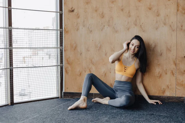 Young attractive fitness girl sitting on the floor near the window on the background of a wooden wall, resting on yoga classes — Stock Photo, Image