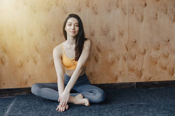 Joven chica atractiva fitness sentada en el suelo cerca de la ventana en el fondo de una pared de madera, descansando en clases de yoga — Foto de Stock