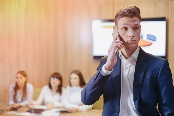 Stylish young businessman wearing a jacket and shirt on the background of a working office with people talking on a mobile phone