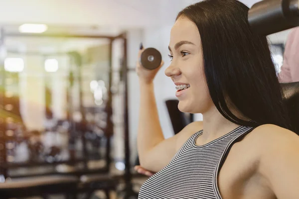 Una joven que usa ropa deportiva en un gimnasio realiza ejercicios de mancuerna, el entrenador la ayuda — Foto de Stock