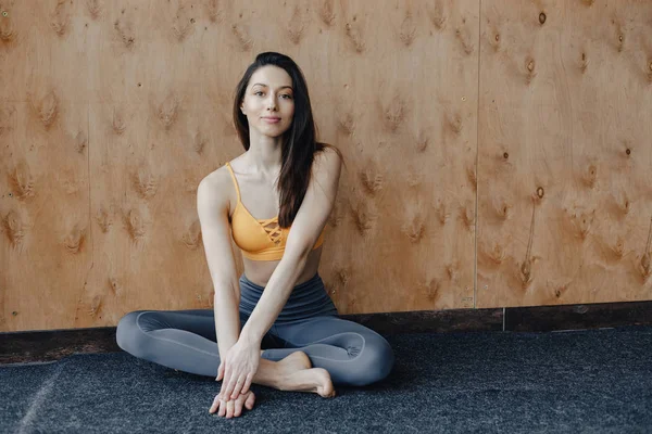 Young attractive fitness girl sitting on the floor near the window on the background of a wooden wall, resting on yoga classes — Stock Photo, Image