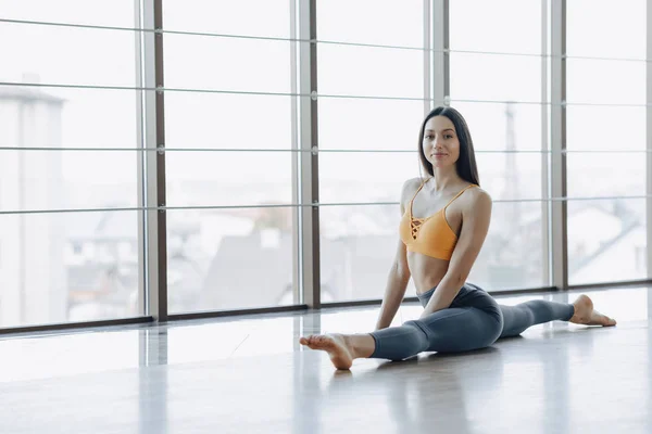 Chica atractiva joven haciendo ejercicios de fitness con yoga en el suelo contra el fondo de ventanas panorámicas — Foto de Stock