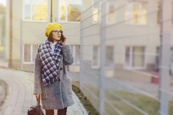 Приваблива молода дівчина в сонцезахисних окулярах в пальто, що йде по вулиці і розмовляє по телефону і посміхається — стокове фото