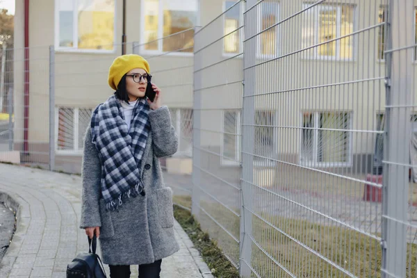 Uma jovem atraente usando óculos de sol em um casaco andando pela rua e falando ao telefone e sorri — Fotografia de Stock