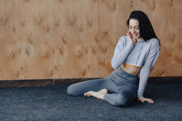 Young attractive fitness girl sitting on the floor near the window on the background of a wooden wall, resting on yoga classes — Stock Photo, Image