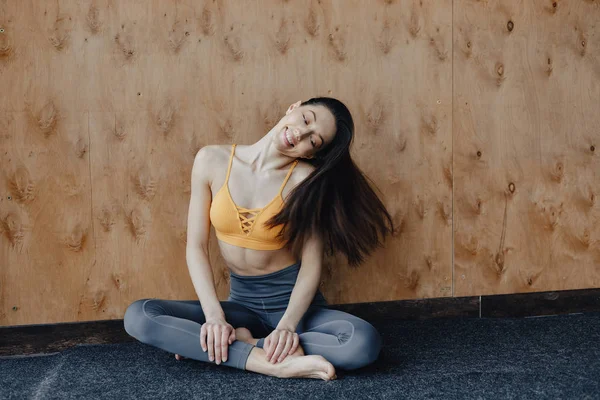 Joven chica atractiva fitness sentada en el suelo cerca de la ventana en el fondo de una pared de madera, descansando en clases de yoga — Foto de Stock