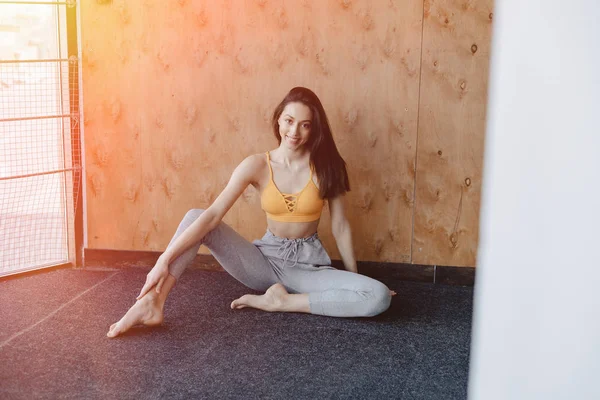 Joven chica atractiva fitness sentada en el suelo cerca de la ventana en el fondo de una pared de madera, descansando en clases de yoga — Foto de Stock