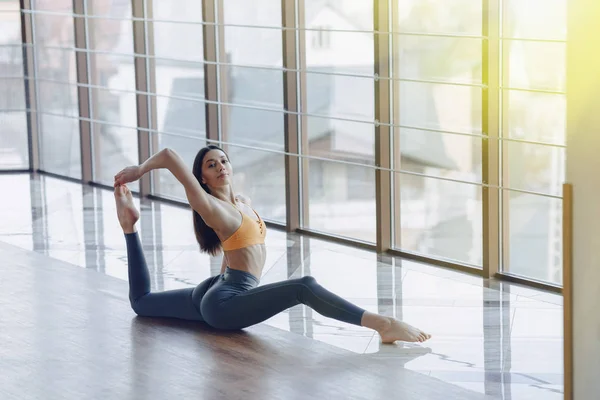 Chica atractiva joven haciendo ejercicios de fitness con yoga en el suelo contra el fondo de ventanas panorámicas — Foto de Stock