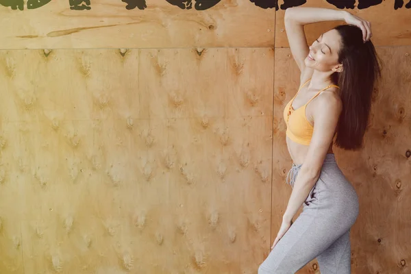 Молода приваблива фітнес-дівчина стоїть біля вікна на фоні дерев'яної стіни, відпочиває на заняттях з йоги — стокове фото