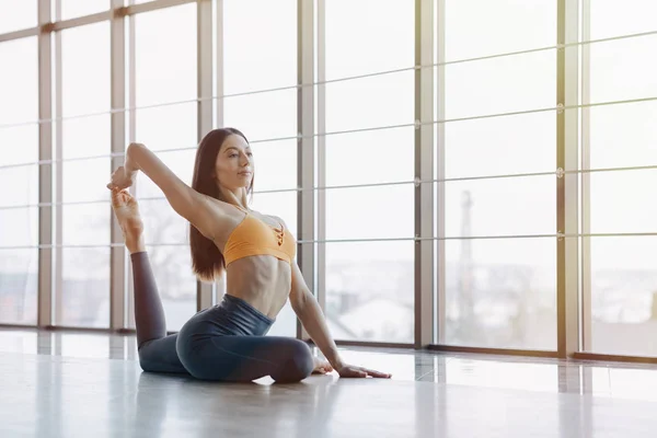 Chica atractiva joven haciendo ejercicios de fitness con yoga en el suelo contra el fondo de ventanas panorámicas — Foto de Stock