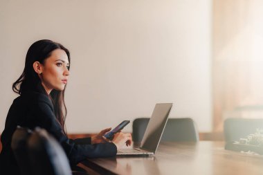 Ofis veya oditoryumda bir dizüstü bilgisayar ve telefon bir masada oturan iş tarzı giysiler genç çekici duygusal kız