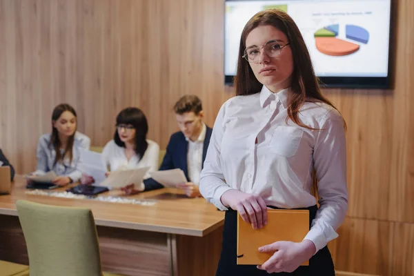 Joven atractiva chica de oficina elegante trabajador en gafas con un cuaderno en las manos en el fondo de los colegas de trabajo — Foto de Stock