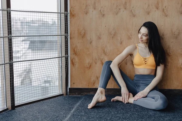 Joven chica atractiva fitness sentada en el suelo cerca de la ventana en el fondo de una pared de madera, descansando en clases de yoga — Foto de Stock