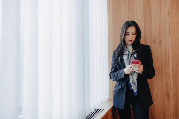 Joven chica atractiva emocional en ropa de negocios en una ventana con un teléfono en una oficina moderna o auditorio — Foto de Stock