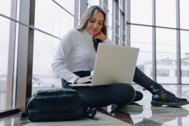 Bilgisayarla çalışan çekici bir kız havaalanı terminalinde ya da ofisin zemininde çalışıyor. seyahat atmosferi ya da alternatif çalışma ortamı. Serbest çalışan öğrenci iş toplantısına gidiyor. Alternatif iş yeri, teknoloji ve bekleme kavramı.