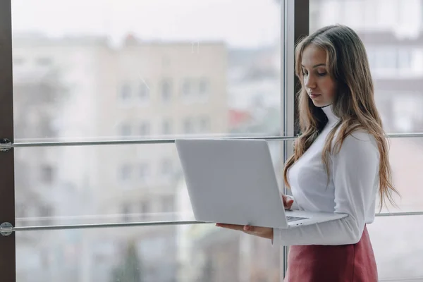 pretty young girl with laptop by the window in office with big windows. remote work concept. freelancer working alone at laptop.