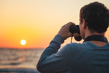 Gün batımında deniz kıyısında elinde dürbünolan bir adam.