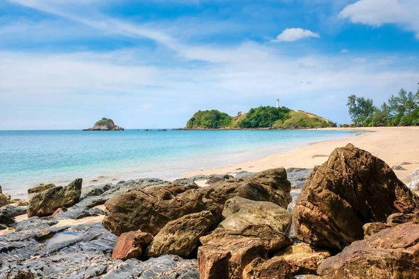 Güzel bir tropikal plaj. Sarı kum ve ön planda büyük taşlar, turkuaz deniz ve bulutlu mavi gökyüzü. Uzaktaki bir uçurumda beyaz bir deniz feneri. Koh Lanta, Tayland.