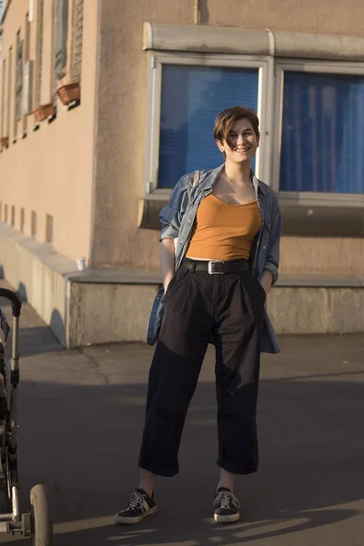 Joven chica de moda en un tema naranja, una camisa de mezclilla y pantalones anchos se encuentra cerca de la casa al atardecer — Foto de Stock