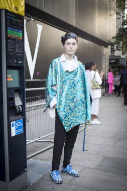 Londra, İngiltere - 14 Eylül 2018: Londra Moda Haftası sırasında sokakta insanlar. Mavi ile beyaz lekeli mavi spor ayakkabı giyen bir panço yazdırma ve siyah deri bere ile genç adam