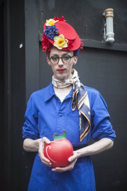 Siyah bir duvara karşı duran bir adam sarı pantolon, mavi bir ceket ve çilek ile kafasına bir çelenk şeklinde kırmızı çanta