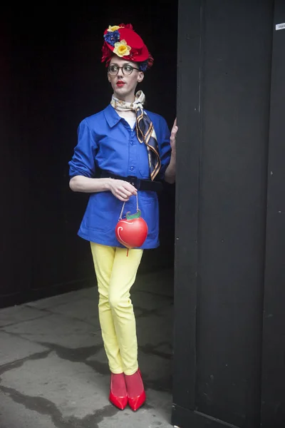 Un hombre con pantalones amarillos, una chaqueta azul y una bolsa roja en forma de fresa con una corona en la cabeza está de pie contra una pared negra. —  Fotos de Stock