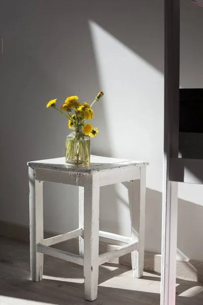 Bouquet di denti di leone gialli in un vaso di vetro trasparente su uno sgabello di legno bianco sullo sfondo della parete — Foto Stock