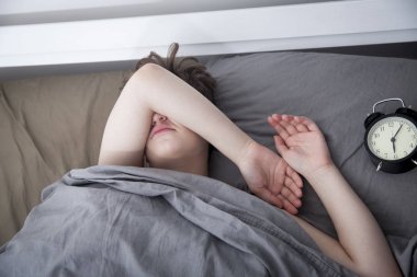 kız, gri bir battaniye sarılmış, alarmı kapatmak için elini uzatır. Çalar saatin altı saati var..