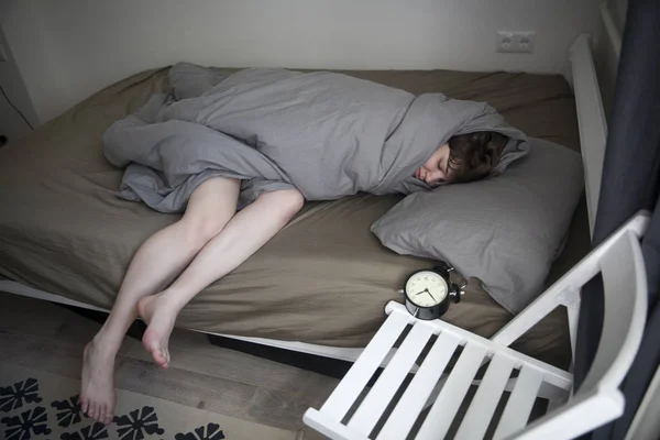 Mädchen, in eine graue Decke gehüllt, streckt ihre Hand aus, um den Wecker auszuschalten. Es stehen sechs Stunden auf dem Wecker. — Stockfoto