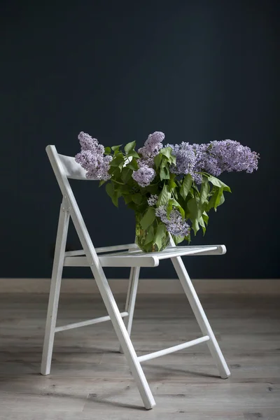 The bouquet of lilac twigs in a transparent green jar on the white chair as a decoration of interior opposite of black wall — Stock Photo, Image