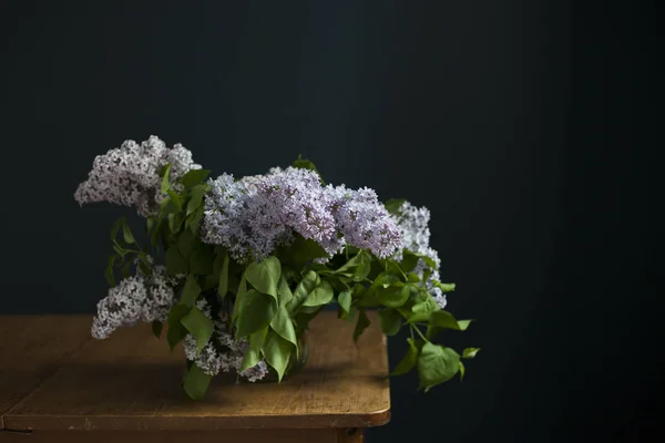 Bouquet of lilac twigs in a transparent green jar on the wooden table opposite of black wall — Stock Photo, Image