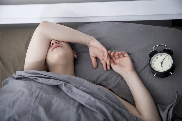 Girl, wrapped in a grey blanket, puts out her hand to turn off the alarm. There is six hours on the alarm clock. — Stock Photo, Image