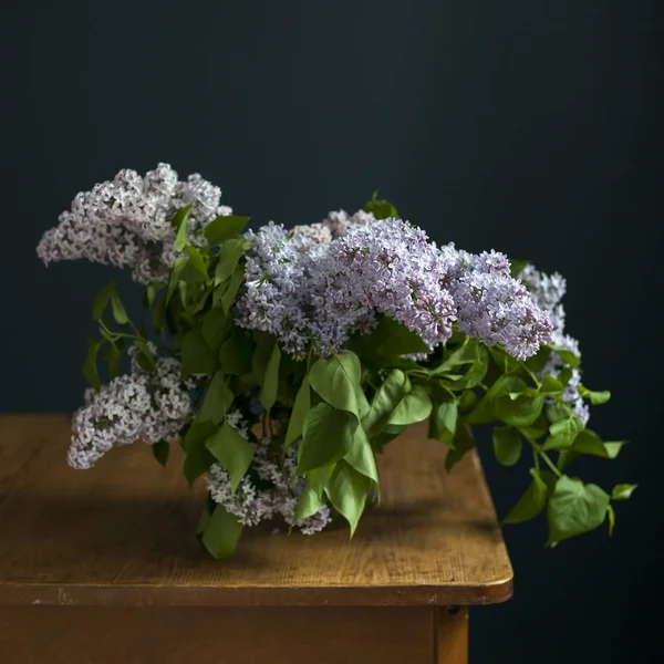 Bouquet of lilac twigs in a transparent green jar on the wooden table opposite of black wall — Stock Photo, Image