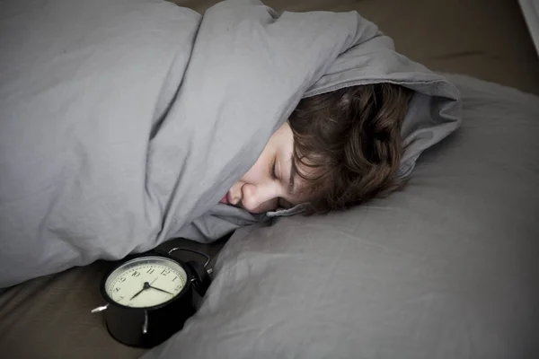 Girl, wrapped in a grey blanket, puts out her hand to turn off the alarm. There is six hours on the alarm clock. — Stock Photo, Image