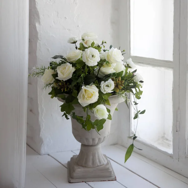 Bouquet of white artificial roses, eucalyptus and ivy in a stone vase on the windowsill near the window — Stock Photo, Image