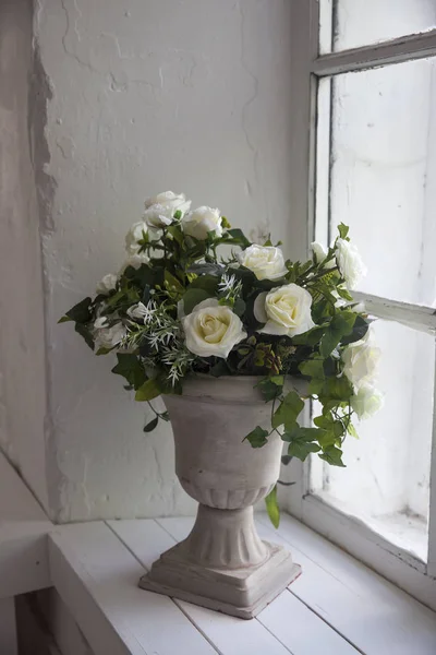 bouquet of white artificial roses, eucalyptus and ivy in a stone vase on the windowsill near the window