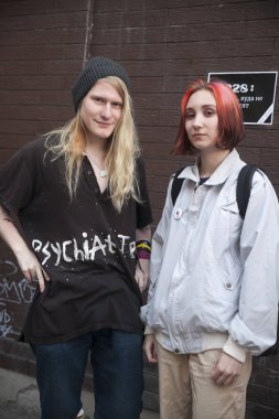 Örme şapkalı uzun saçlı, siyah tişörtlü ve kot pantolonlu genç sarışın bir adam kot ceketli kırmızı saçlı kız arkadaşının yanında duruyor.