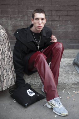 Siyah ceketli, bordo deri pantolonlu ve yüzünde çapraz şekilli dövmeli genç bir adam kaldırımda oturuyor.