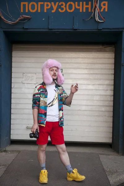 Un joven con una camisa variopinta de estilo hawaiano, una camiseta blanca, pantalones cortos rojos y un sombrero de piel rosa con una cámara se para frente a la salida del garaje. — Foto de Stock