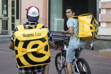 Food delivery in Moscow - courier in a hat with earflaps and a yellow jacket with inscription Yandex food and yellow backpack rides on bicycle on street clipart