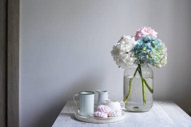 Bir buket çok renkli ortanca, beyaz bir arka planda 5 litrelik kavanozda. Kupalı bir tepsi, sütçü ve pembe şekerleme. Boşluğu kopyala