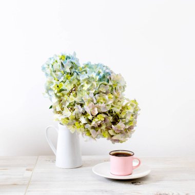Beyaz sürahi içinde yeşil ve mavi pastel ortancayla bej masa üzerinde büyük bir dal. Pembe bir sürahi içinde yeşil ve mavi pastel ortancalı büyük bir dal bej masa üzerinde bir fincan kahve ve süt. Boşluğu kopyala