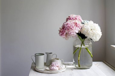 Bir buket çok renkli ortanca, beyaz bir arka planda 5 litrelik kavanozda. Kupalı bir tepsi, sütçü ve pembe şekerleme. Boşluğu kopyala