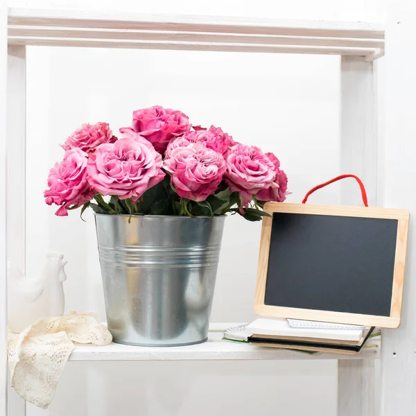 Bouquet of red roses in a tin bucket on a white wall background. Black slate for writing on a stack of notebooks on a rack. Copy space