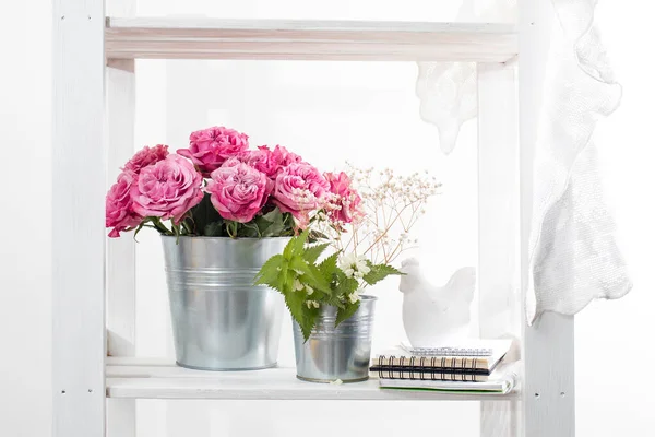 Bouquet of red roses in a tin bucket on a white wall background. Wild white dead-nettle grass in a tin can and porcelain chicken figurine on notebooks and lace fabric. Copy space