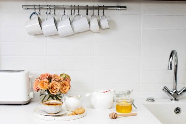 Düğün buketi pembe ve turuncu lisianthus, antirrhinum ve okaliptüs mutfak masasında cam vazoda. Beyaz fincanda siyah çay, bal kavanozu ve bal kaşığı, beyaz mutfakta limon kaplaması.